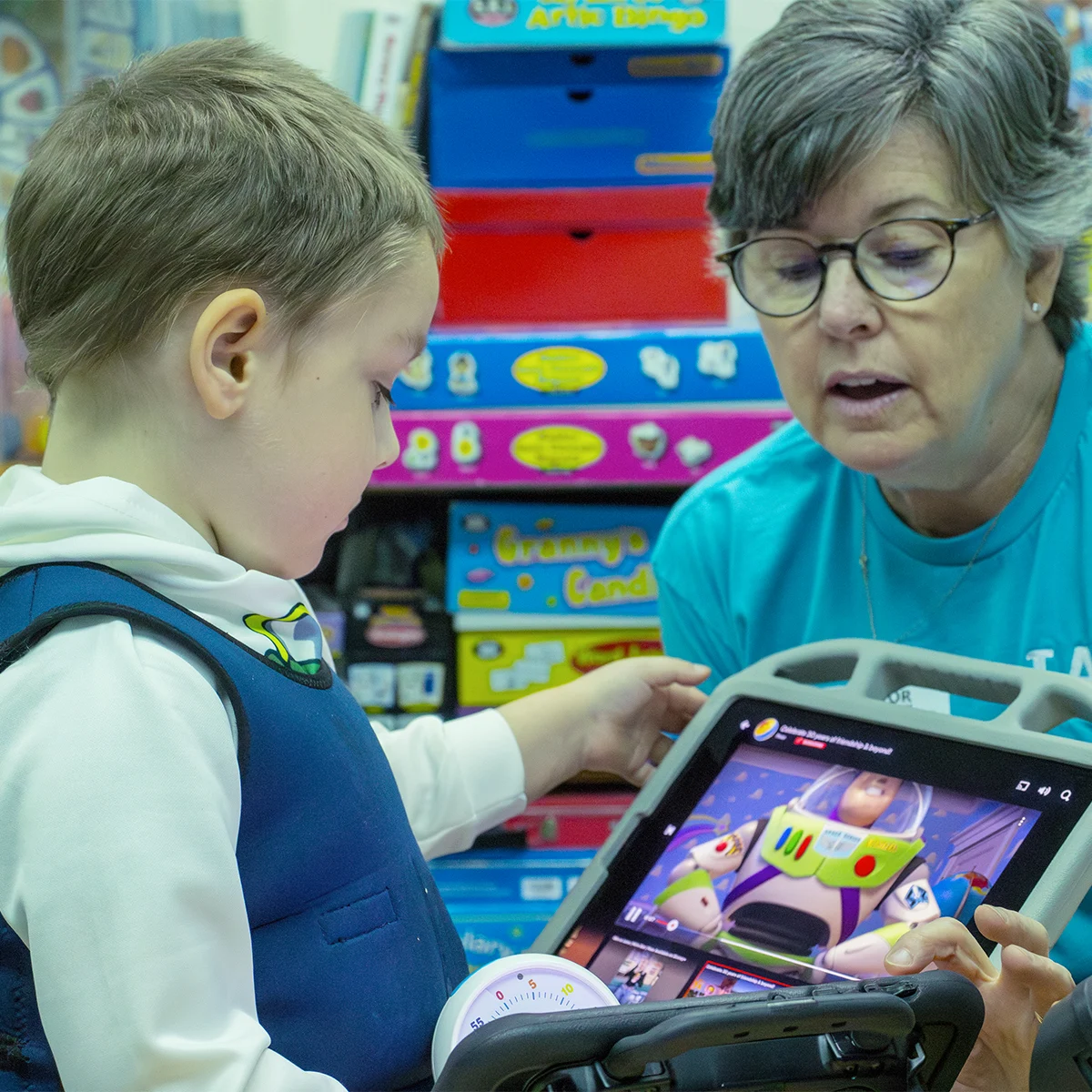 A child and an older person look at a digital tablet displaying Buzz Lightyear from Toy Story, surrounded by board games