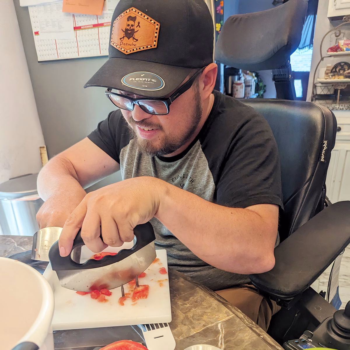 A man in a power wheelchair chopping tomatoes on a cutting board using a U shaped kitchen tool A man in a power wheelchair chopping tomatoes on a cutting board using a U shaped kitchen tool