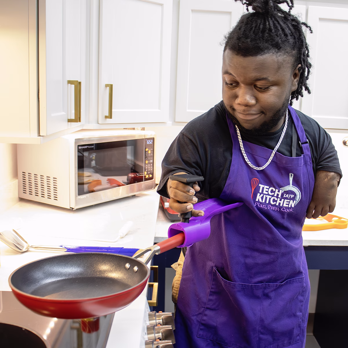 A man in a purple apron using adaptive D printed equipment to hold a frying pan in a kitchen A man in a purple apron using adaptive D printed equipment to hold a frying pan in a kitchen