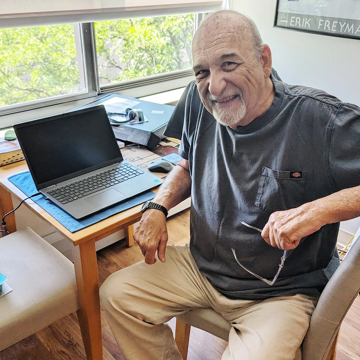 A man sitting at a table with a laptop, holding glasses, in a sunny office space, smiling for the camera