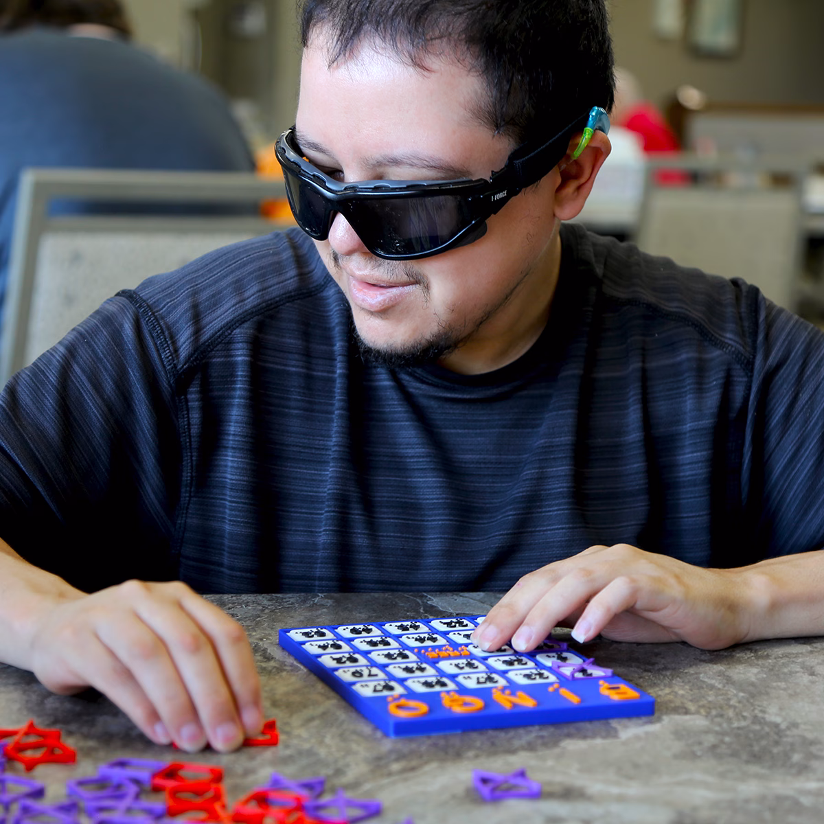 A man wearing protective eyewear plays a tactile bingo game with colorful pieces on a table A man wearing protective eyewear plays a tactile bingo game with colorful pieces on a table
