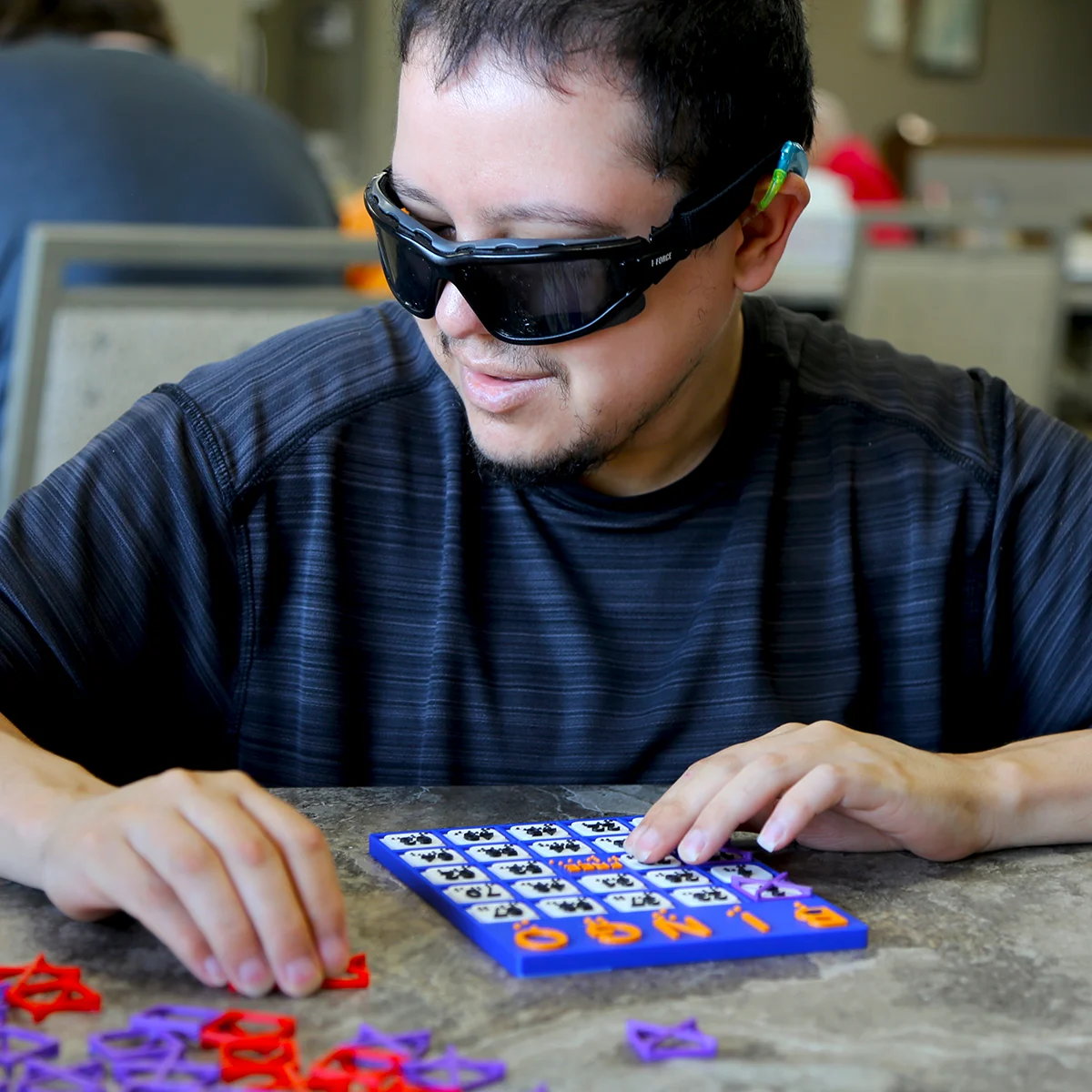 A man wearing protective eyewear plays a tactile bingo game with colorful pieces on a table