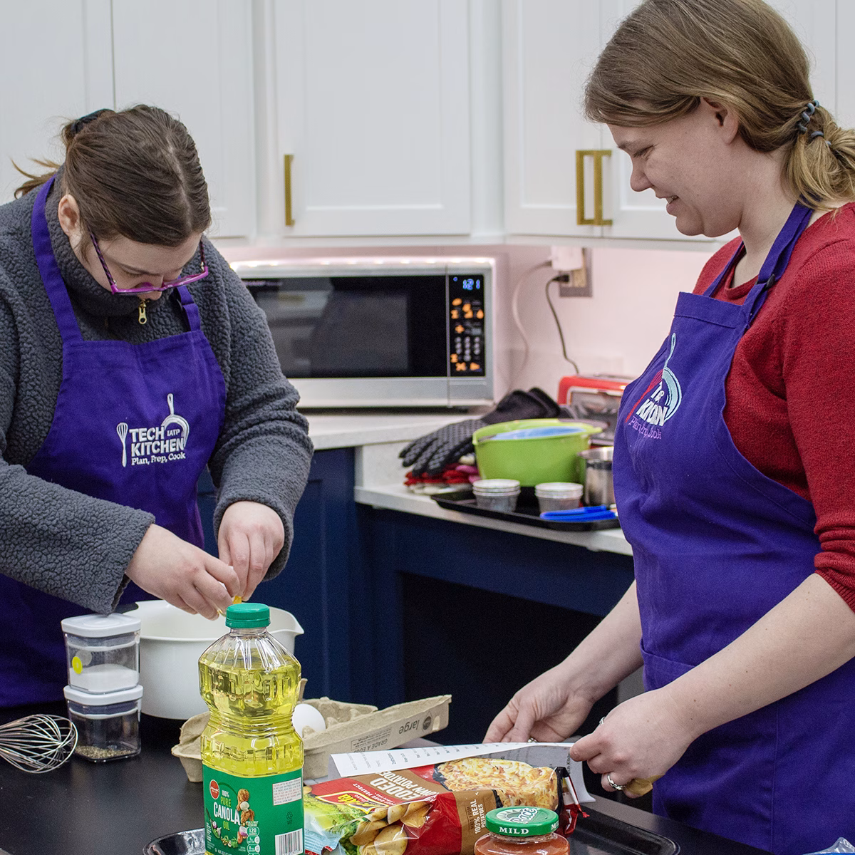 A person in a kitchen preparing food on a table while an instructor stands next to her watching A person in a kitchen preparing food on a table while an instructor stands next to her watching
