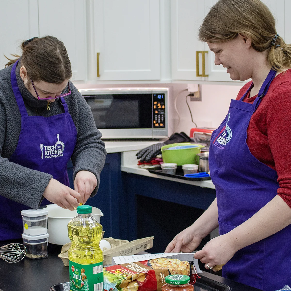 A person in a kitchen preparing food on a table while an instructor stands next to her watching