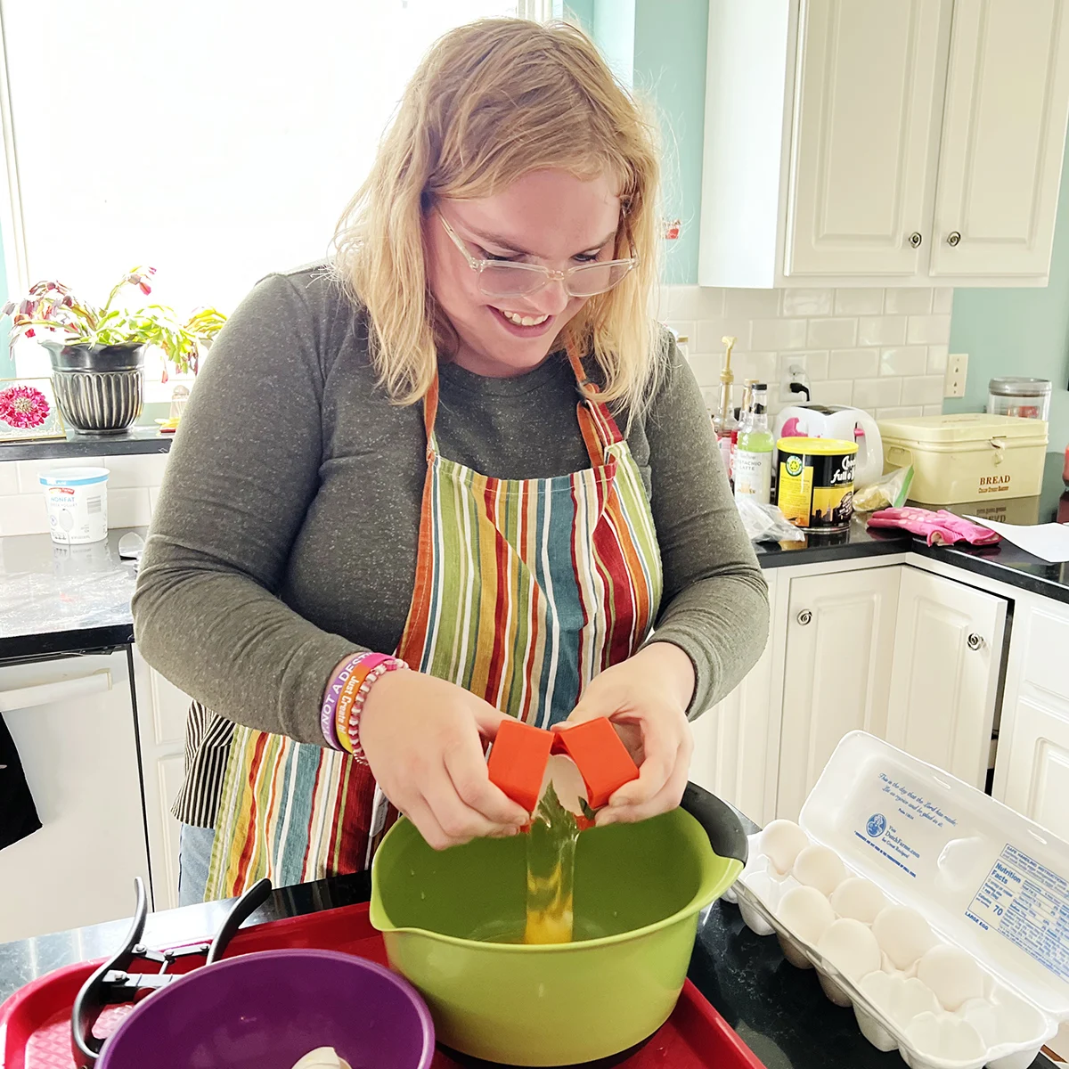 A person in a striped apron cracking an egg with an egg separator into a green bowl in a kitchen