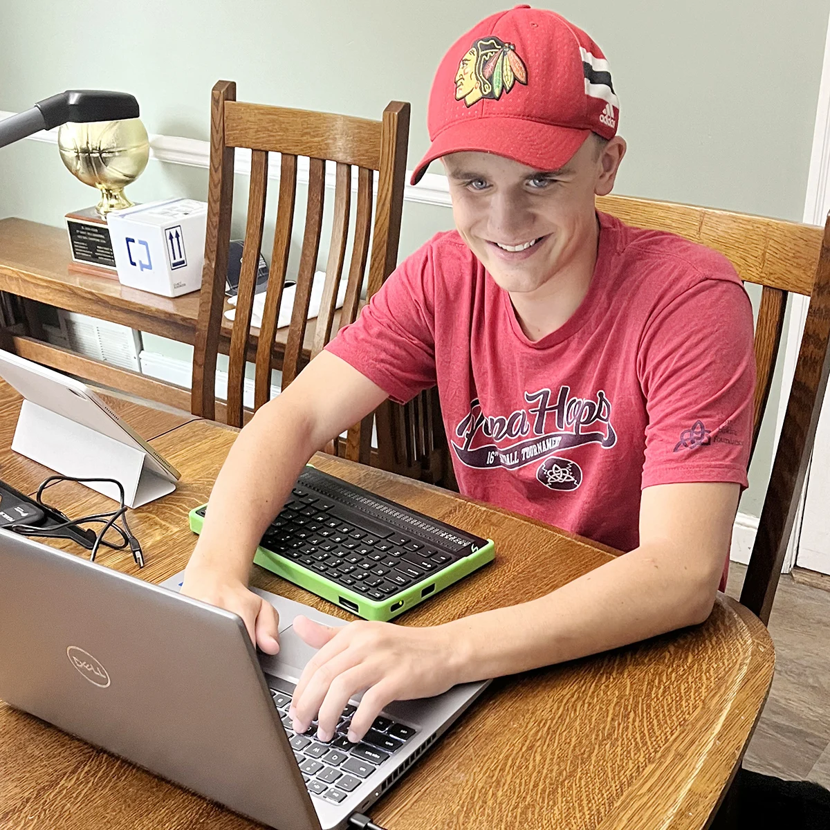 A teen boy in a red cap and T shirt using a laptop with a braille keyboard and display