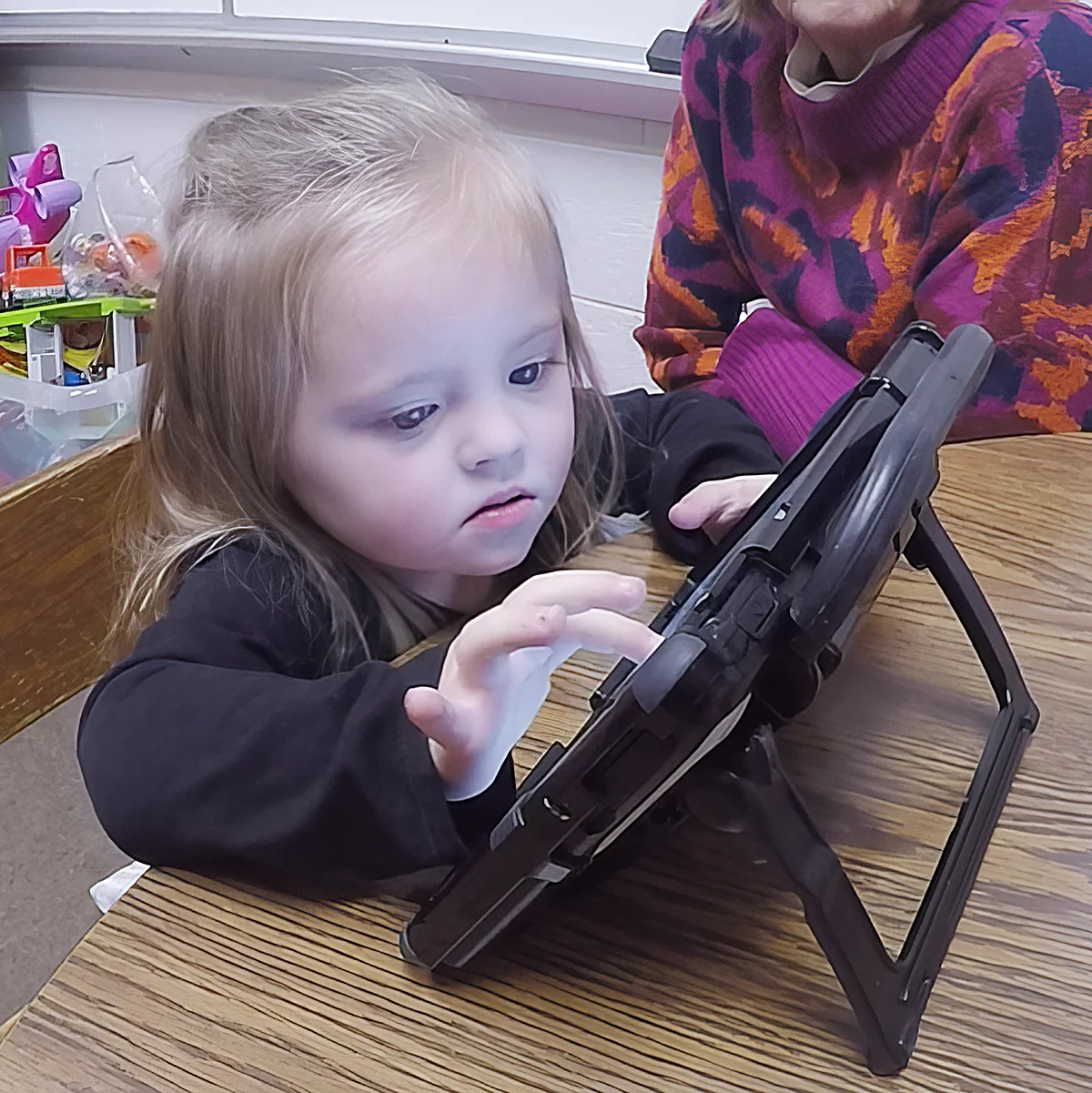 A young child, focused intently on a AAC device resting on a table. The child is tapping on the screen of the device