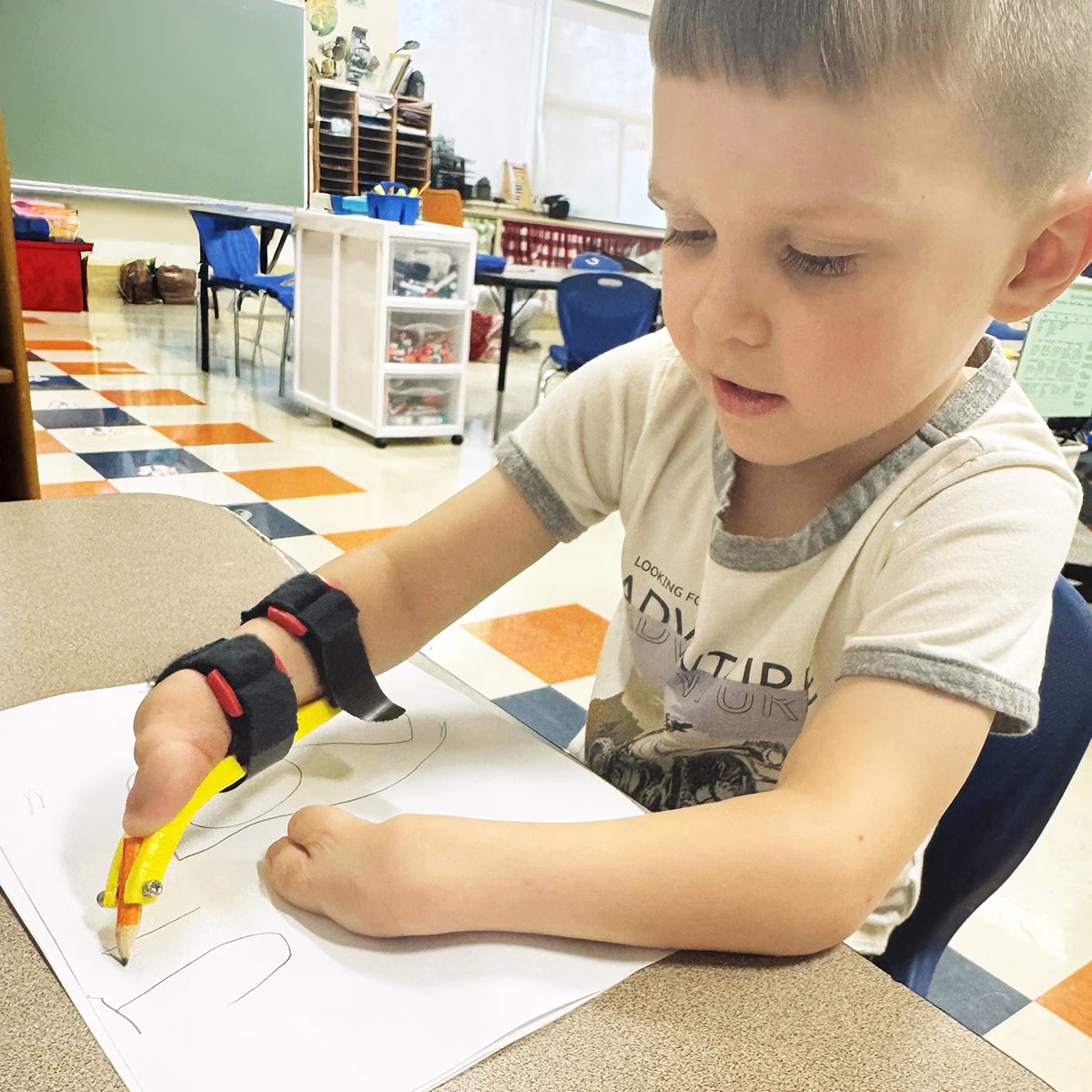 A young child with an adaptive writing aid secured to his right arm with Velcro straps in writing is name, in a classroom setting A young child with an adaptive writing aid secured to his right arm with Velcro straps in writing is name, in a classroom setting