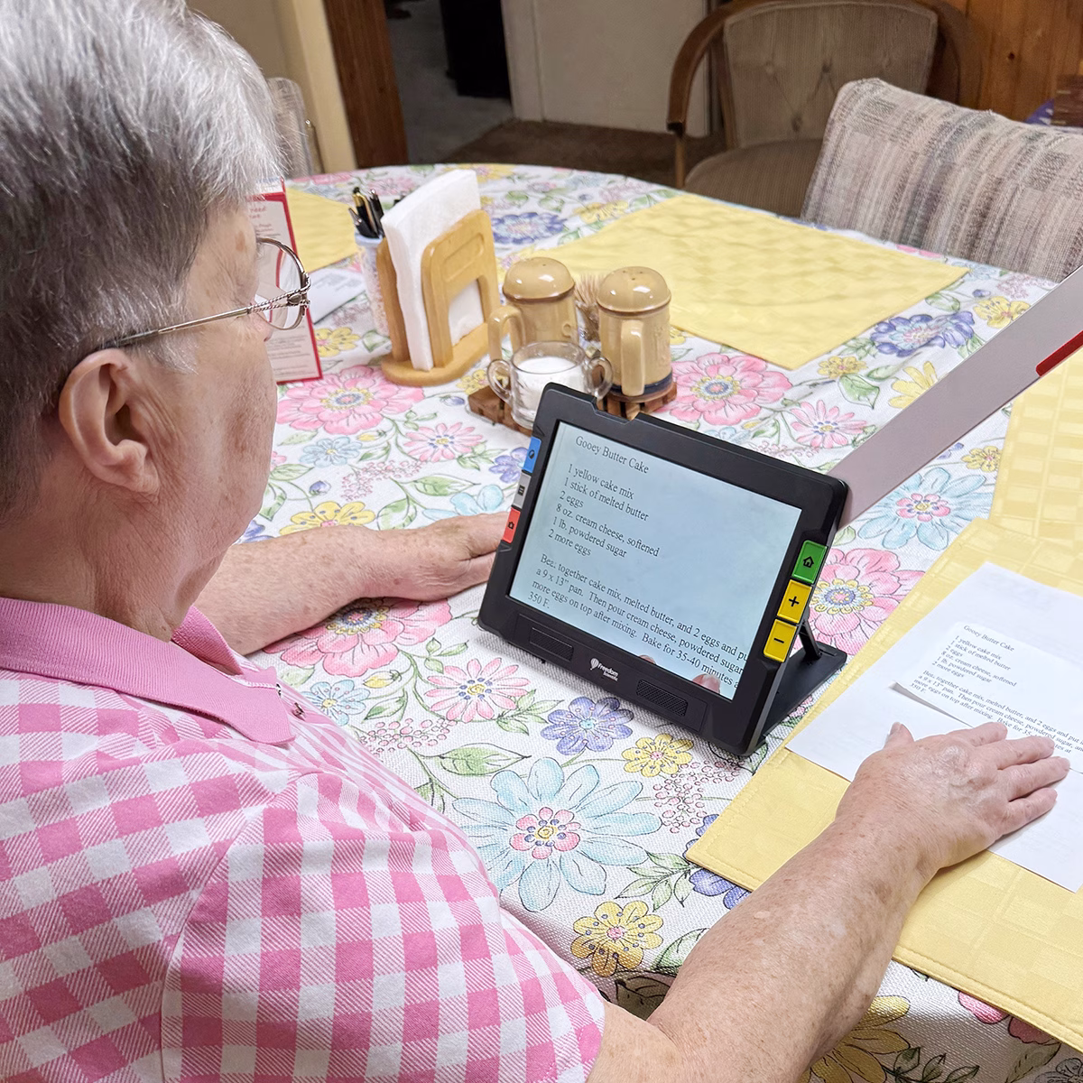 An elderly person reads a recipe on a Ruby device with the extended are out to read a recipe card laying on the table covered with a floral tablecloth An elderly person reads a recipe on a Ruby device with the extended are out to read a recipe card laying on the table covered with a floral tablecloth