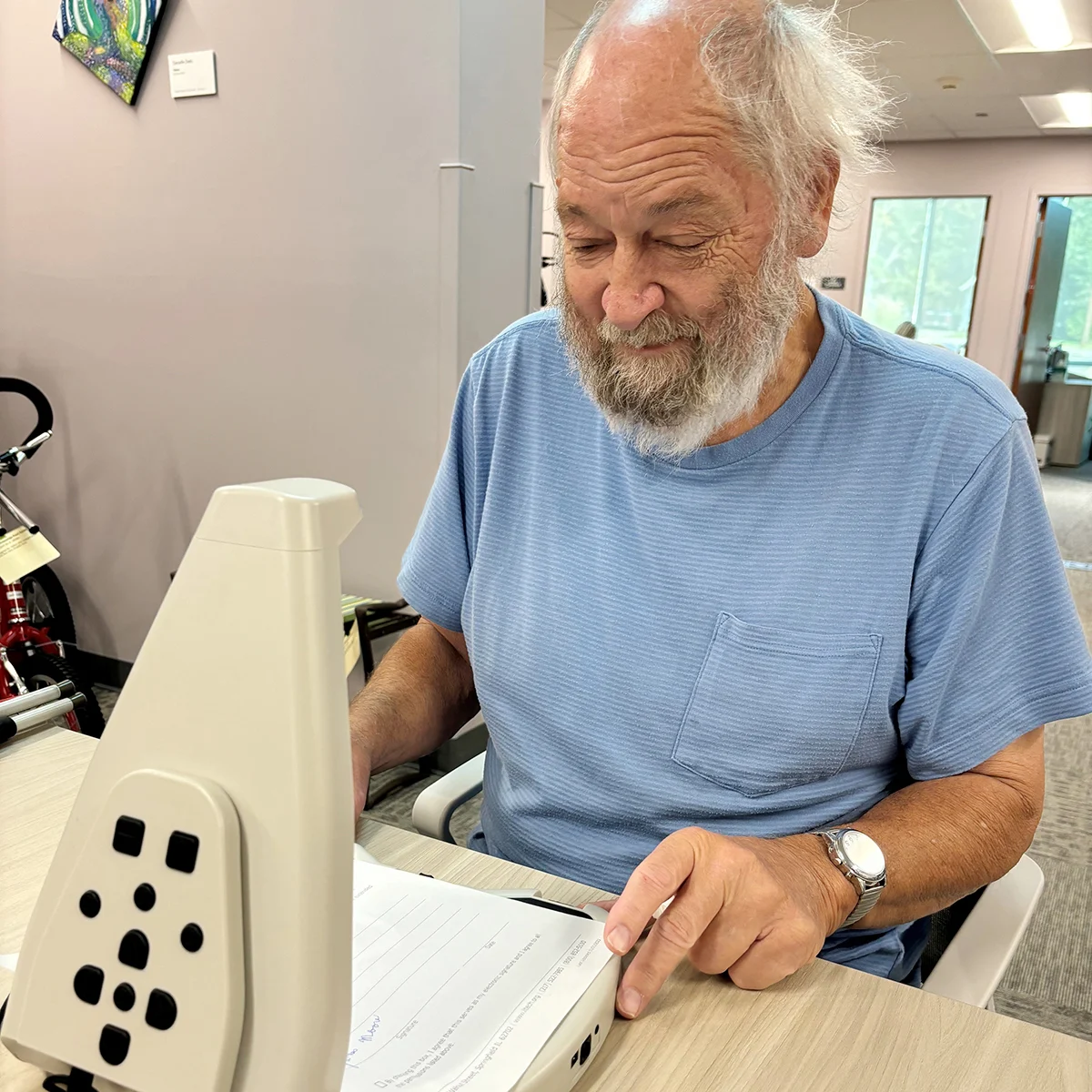Elderly man sitting at a table using LyriQ assistive reader device to read a form