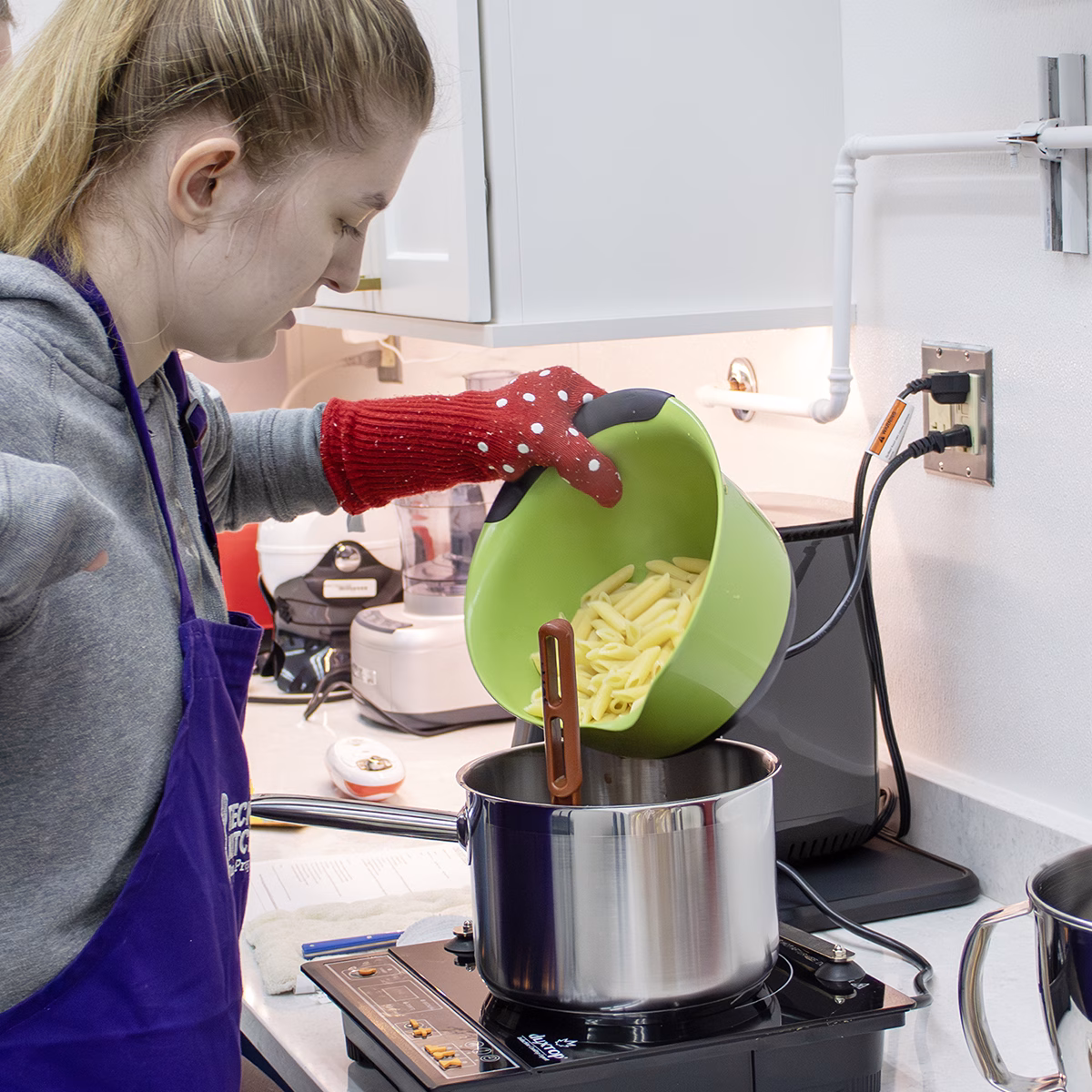 Person pouring pasta from a green bowl into a pot on an induction burner, wearing a purple apron and red oven mitt Person pouring pasta from a green bowl into a pot on an induction burner, wearing a purple apron and red oven mitt