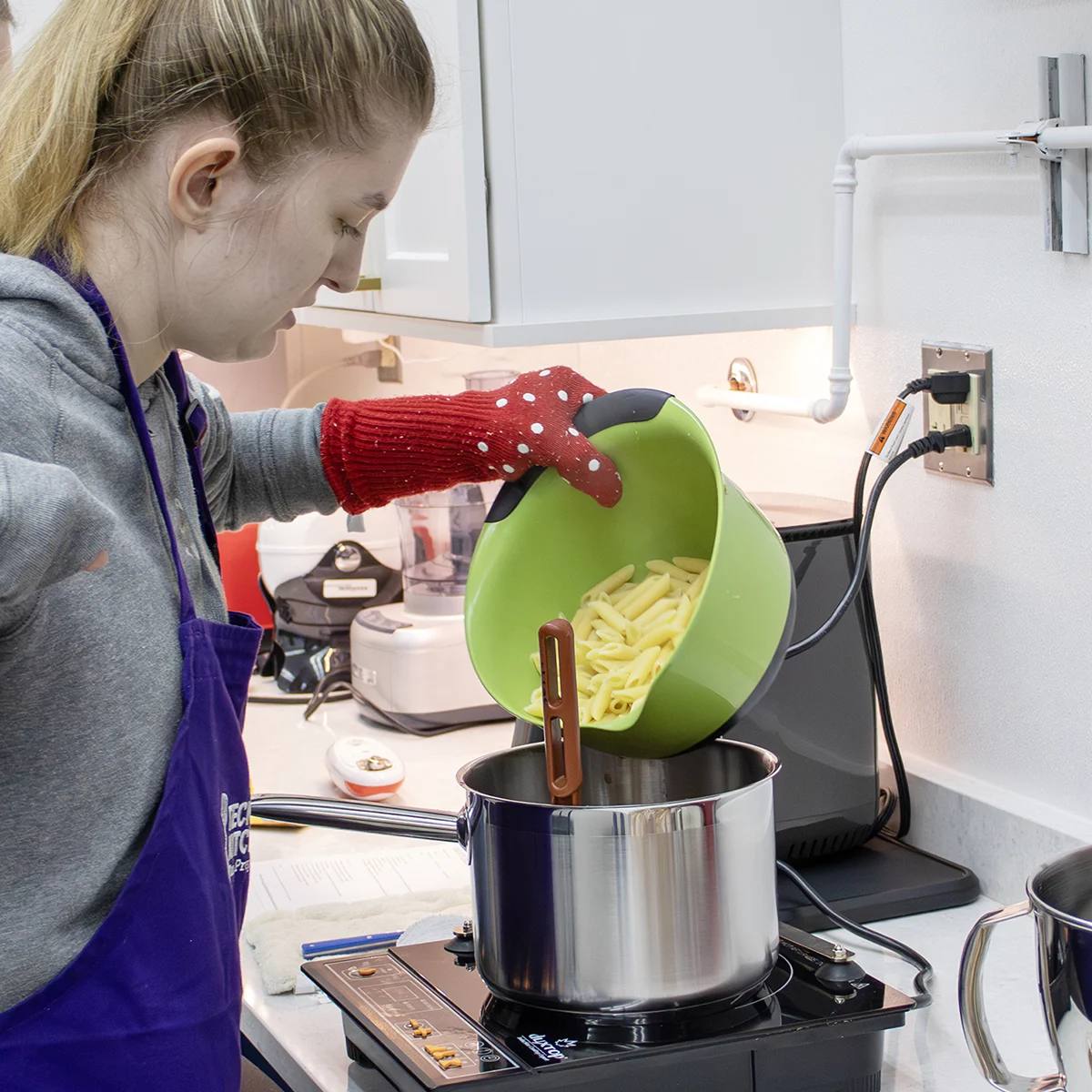 Person pouring pasta from a green bowl into a pot on an induction burner, wearing a purple apron and red oven mitt