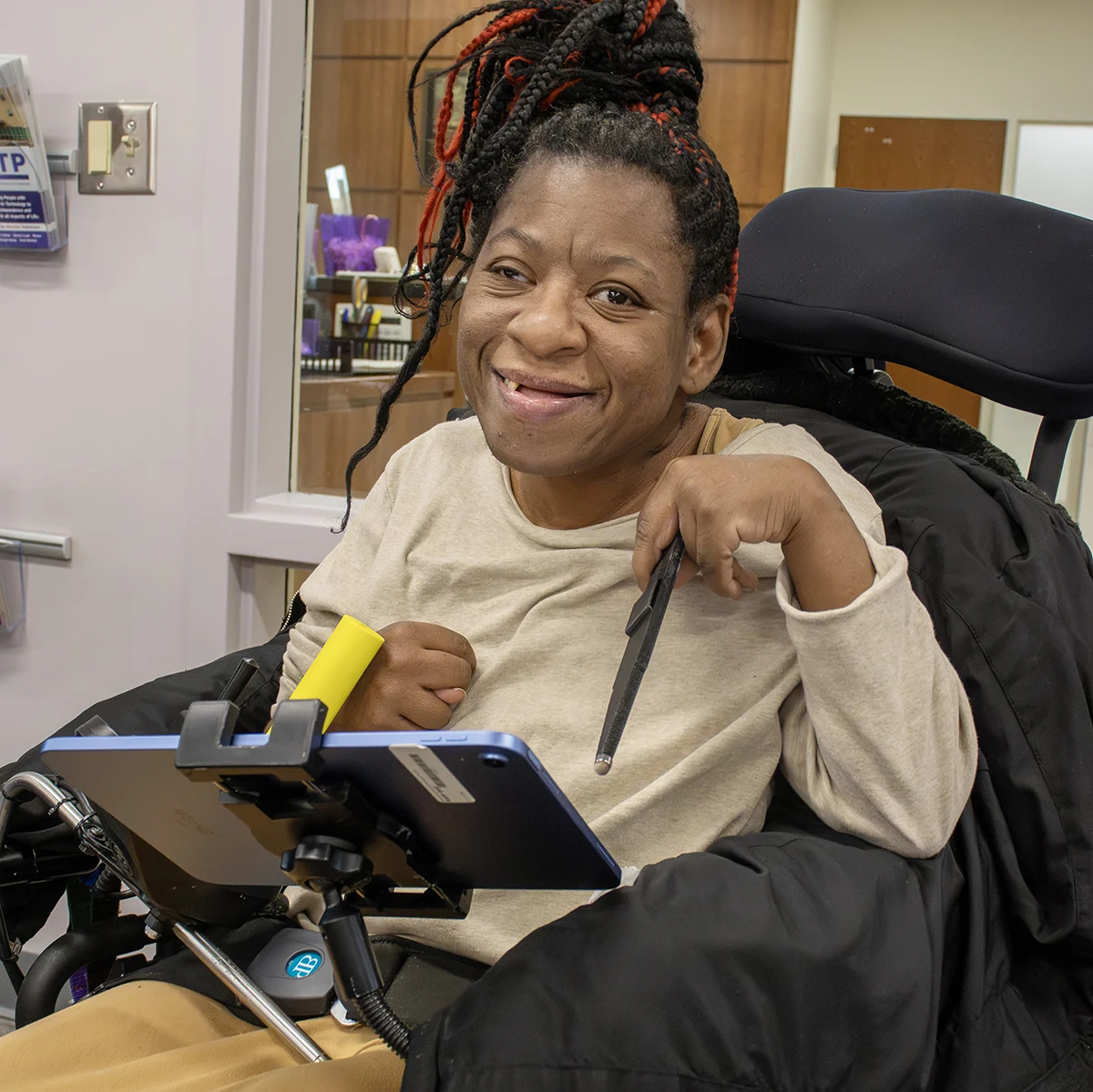 Person smiling in a wheelchair holding a stylus, with a mounted tablet in front of them