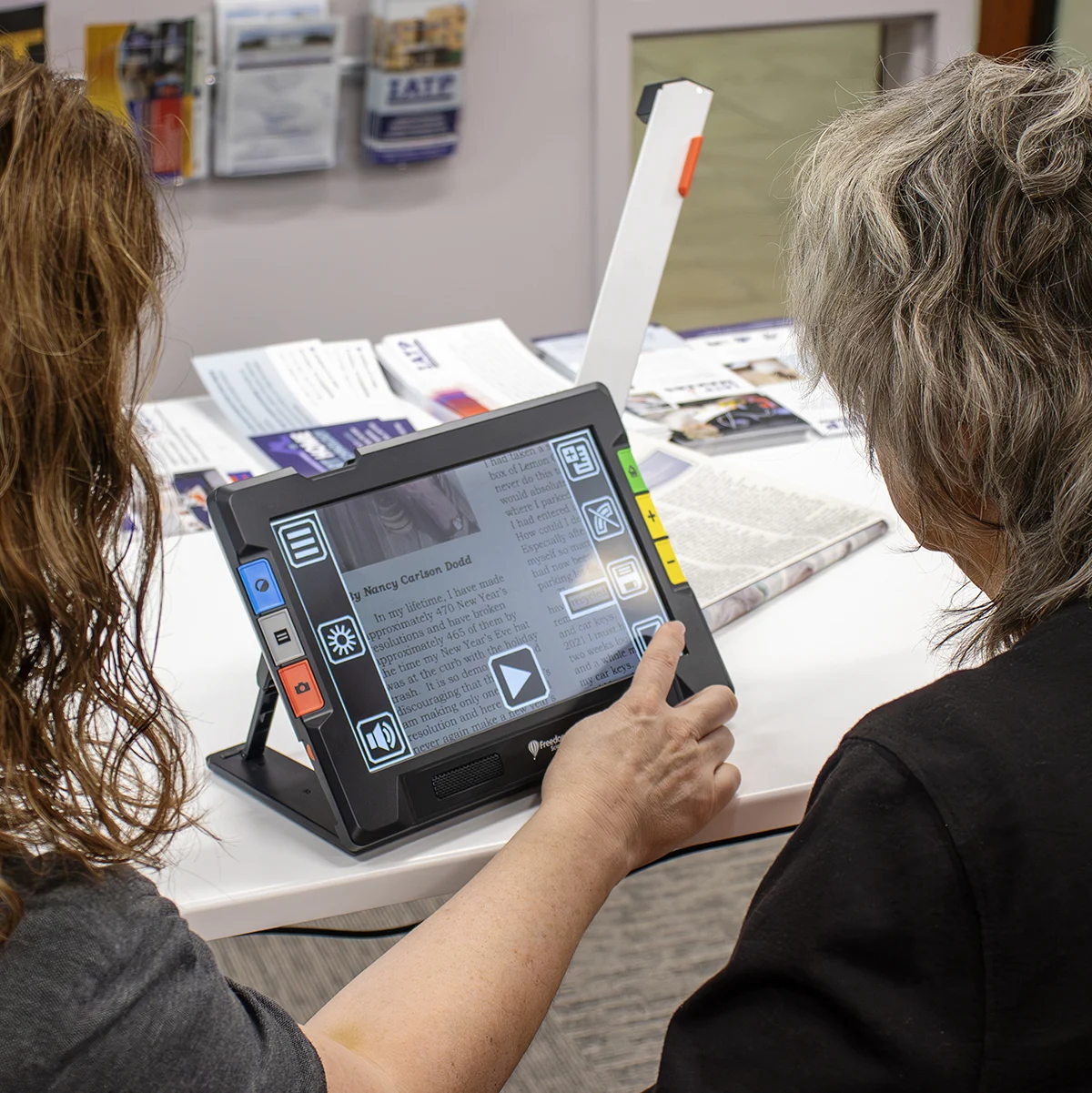 Two people using a magnifier reading device on a table with pamphlets in the background