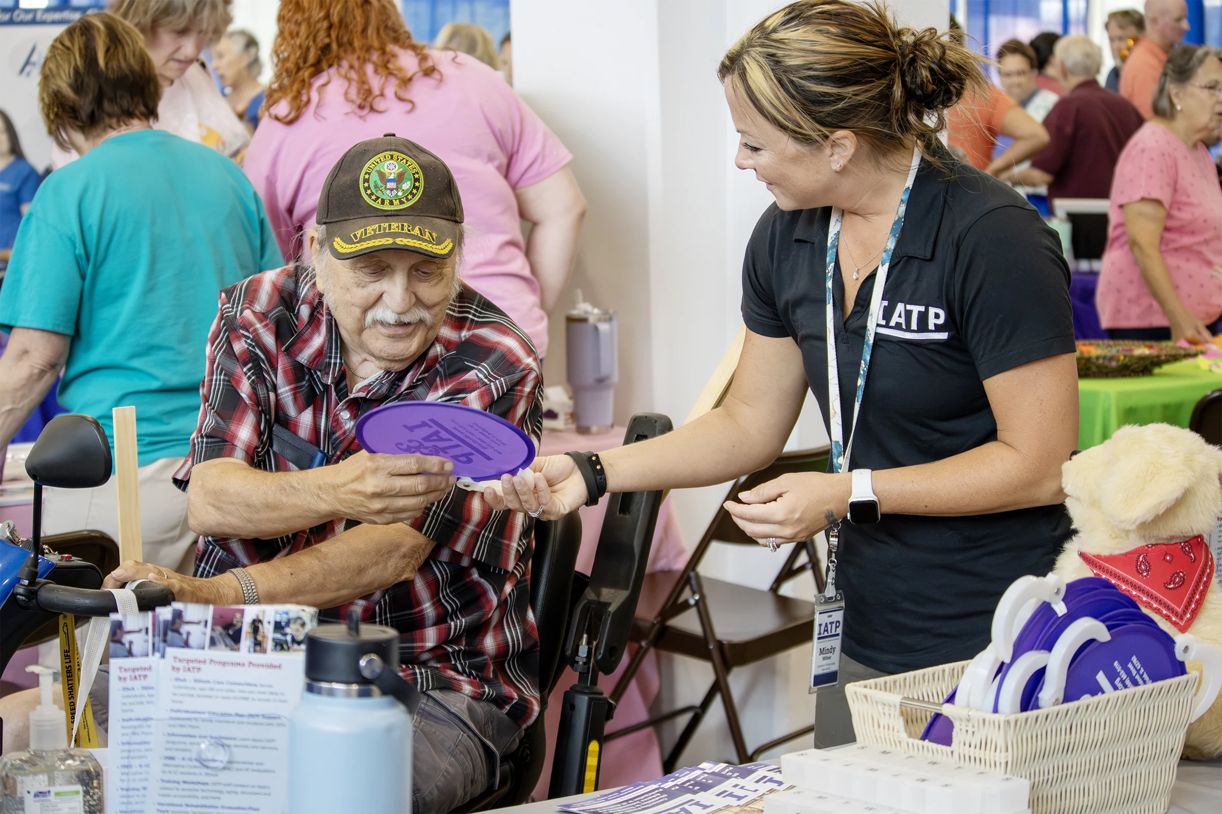 An IATP employee hands a purple fan to an elderly man in a mobility scooter at a community event.