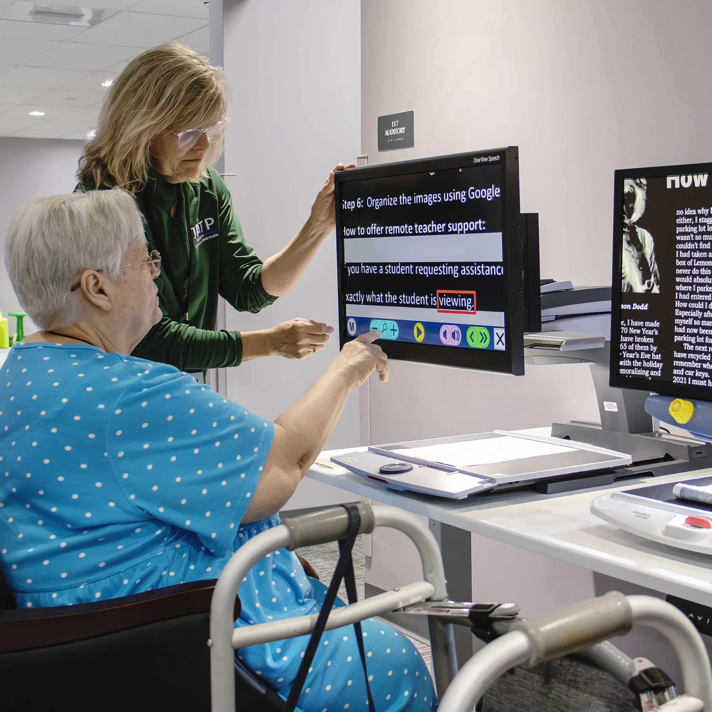 a person sitting looking at a vision magnifier while an IATP staff member stands behind helping to adjust settings in IATP’s Demonstration Center.