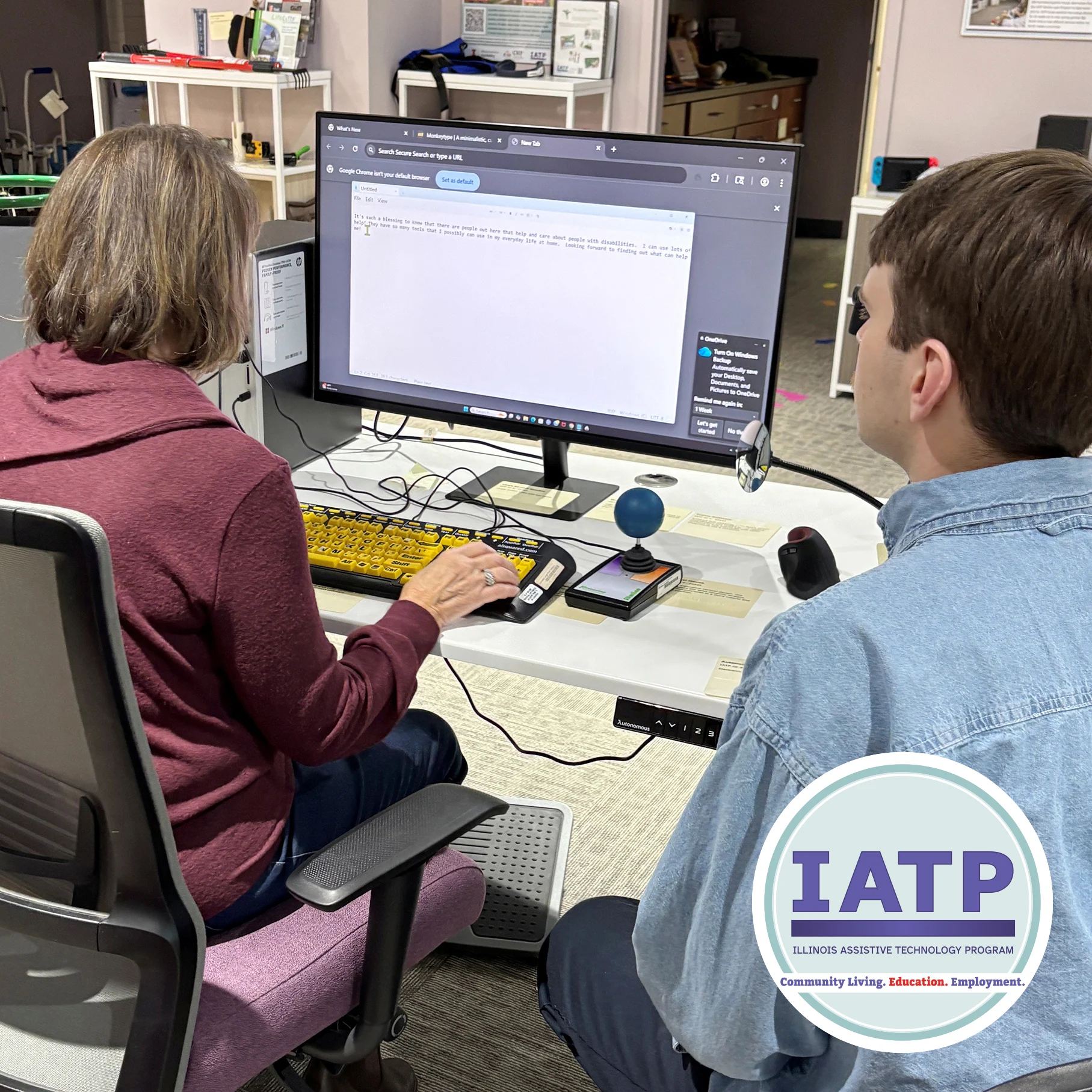 Demo Story Jan B Jan sitting at a computer station in the IATP demonstration center while an IATP staff member looks on. IATP logo sits at the bottom of the photo with the words Community Living, Education and employment under it.