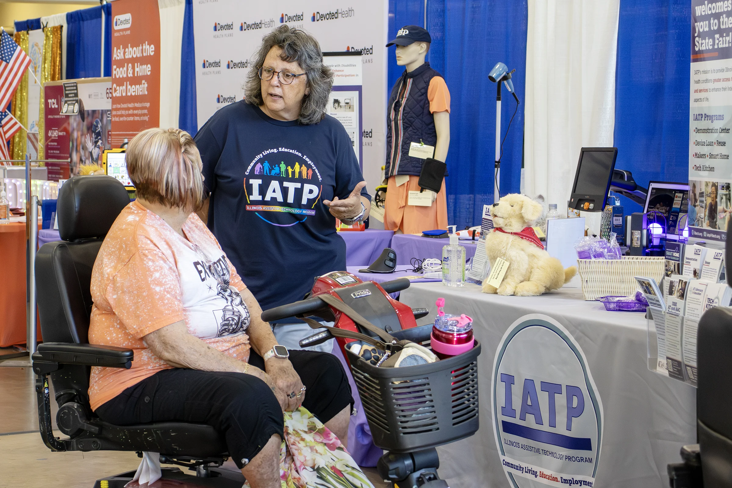 A woman in a motorized wheelchair talks to an IATP staff member at the fair. Various assistive technology items are on display in the background. A woman in a motorized wheelchair talks to an IATP staff member at the fair. Various assistive technology items are on display in the background.
