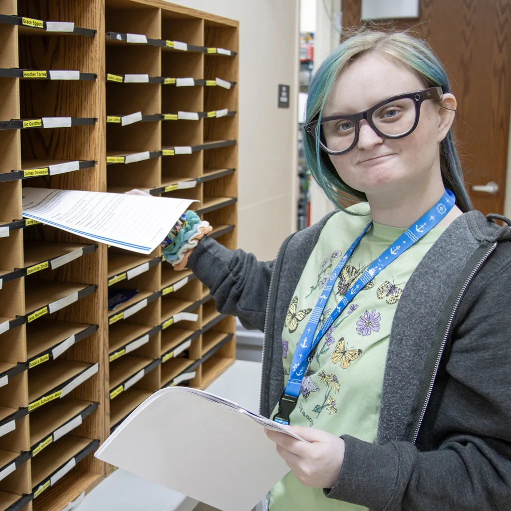 young adult smiling at glasses while standing in front of employee mailboxes at her employer's office