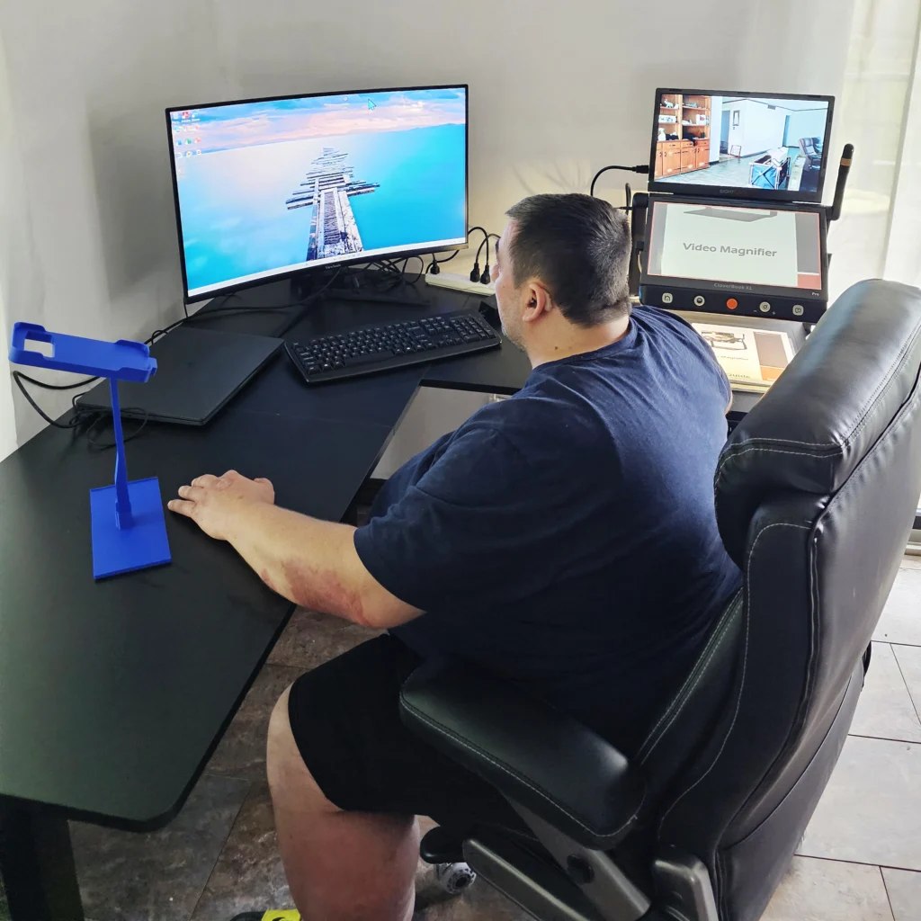 Paul sitting at his home work station with a computer and magnifier in front of him