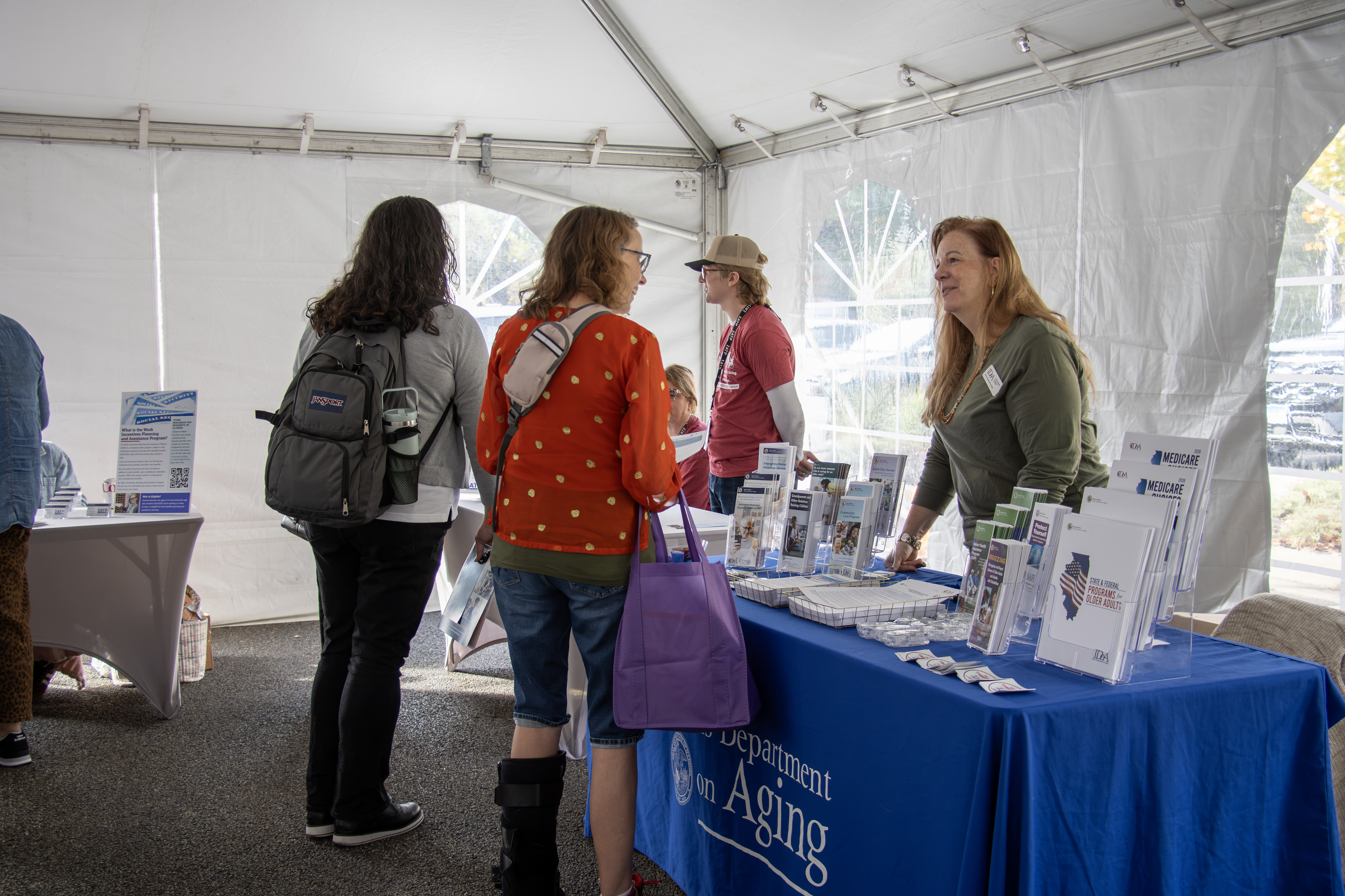 People browse informational tables at the IATP resource booth inside an outdoor event tent