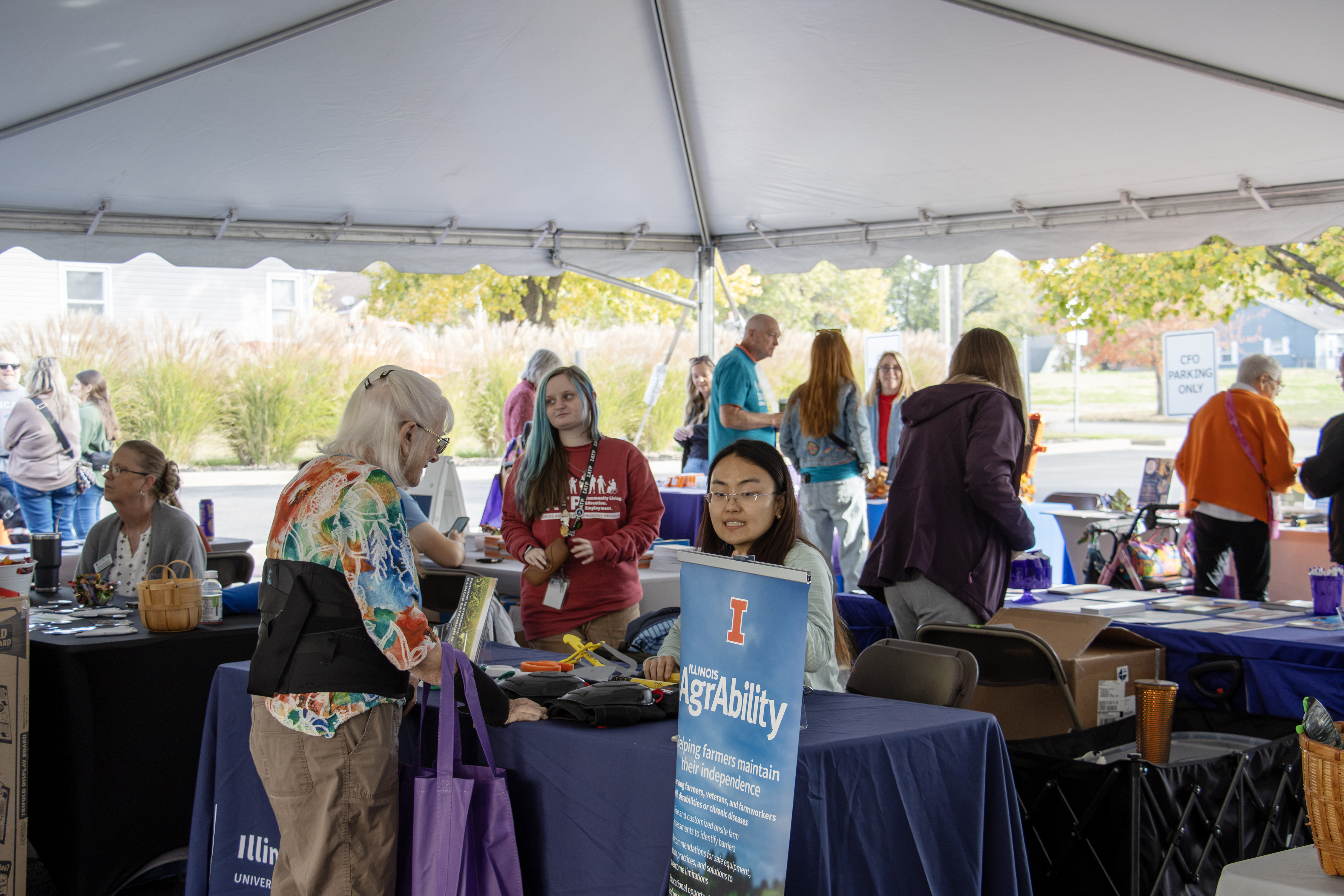 People gather at information booths during an outdoor event beneath a tent, with an Illinois AgrAbility sign in the foreground