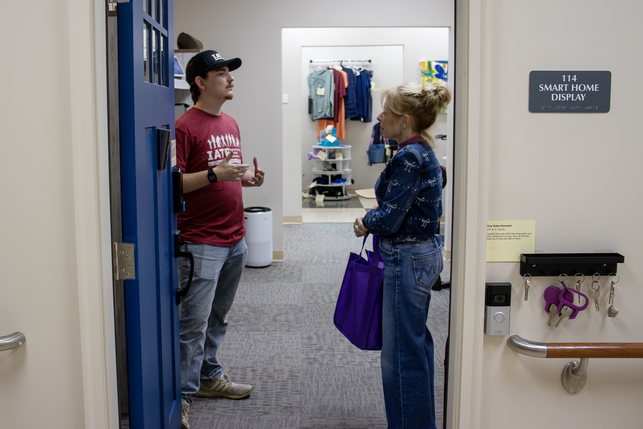 Two adults talk in the doorway of the IATP smart home display, with accessibility devices and a key rack visible on the wall