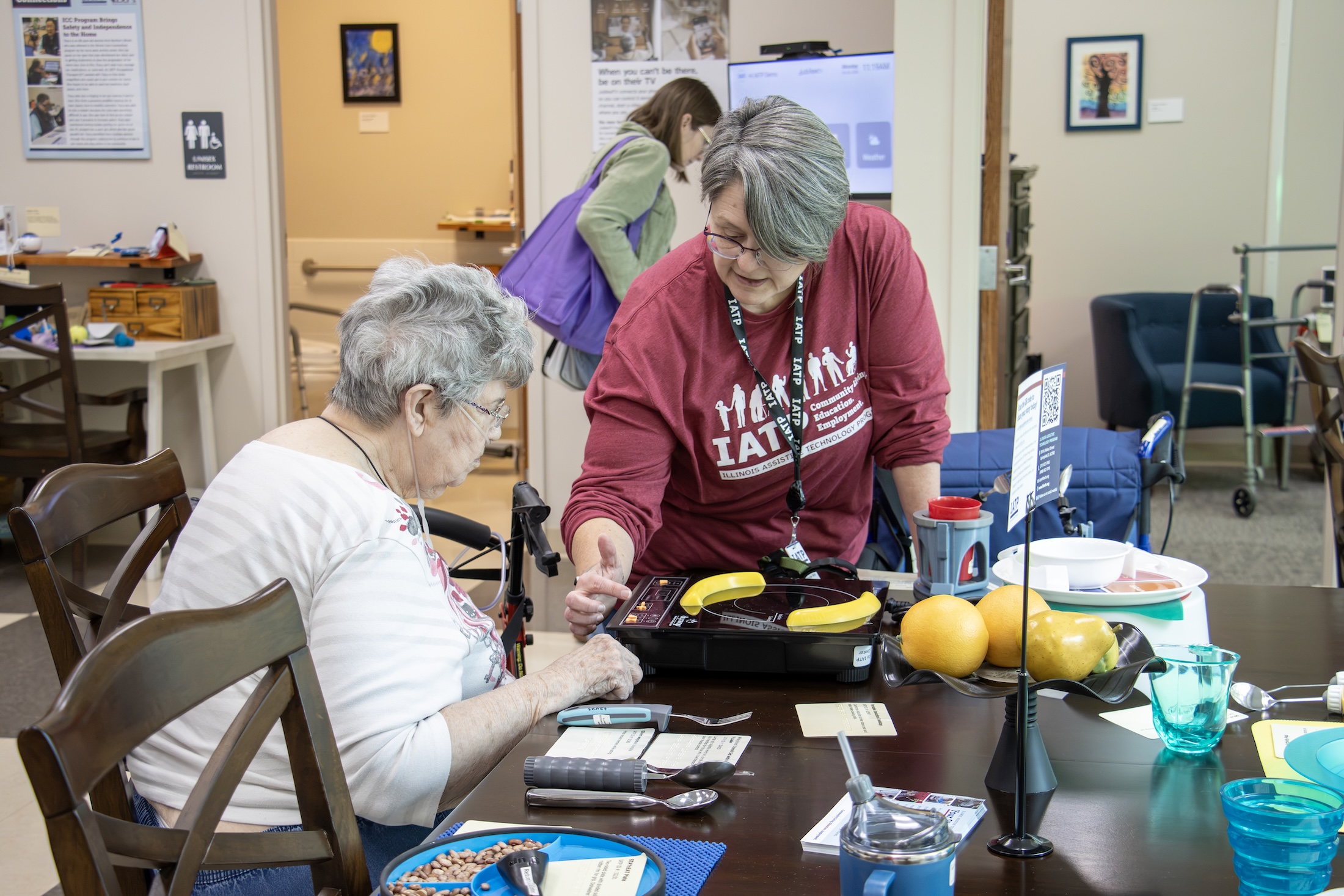 Woman demonstrating an assistive technology device to an older woman seated at a dining table